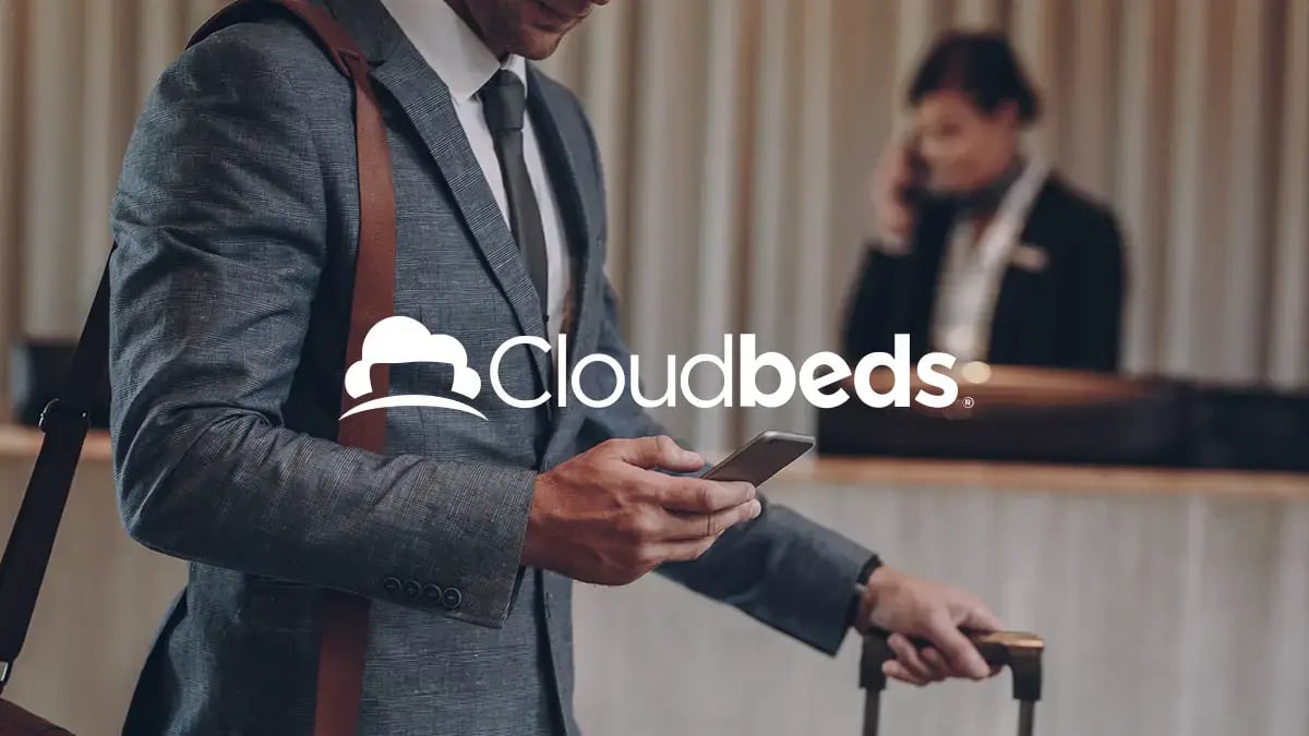 A main in a suit holding his phone and a suitcase at a hotel's desk with Cloudbeds logo in white as an overlay.