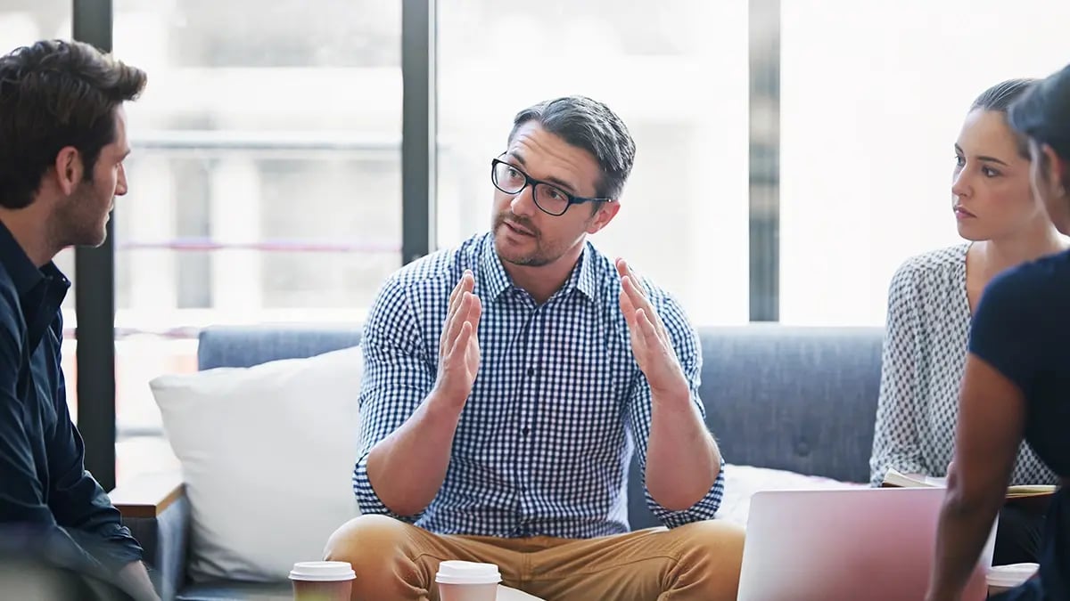 A person in a checked shirt talking to a group of people while sat on a sofa.