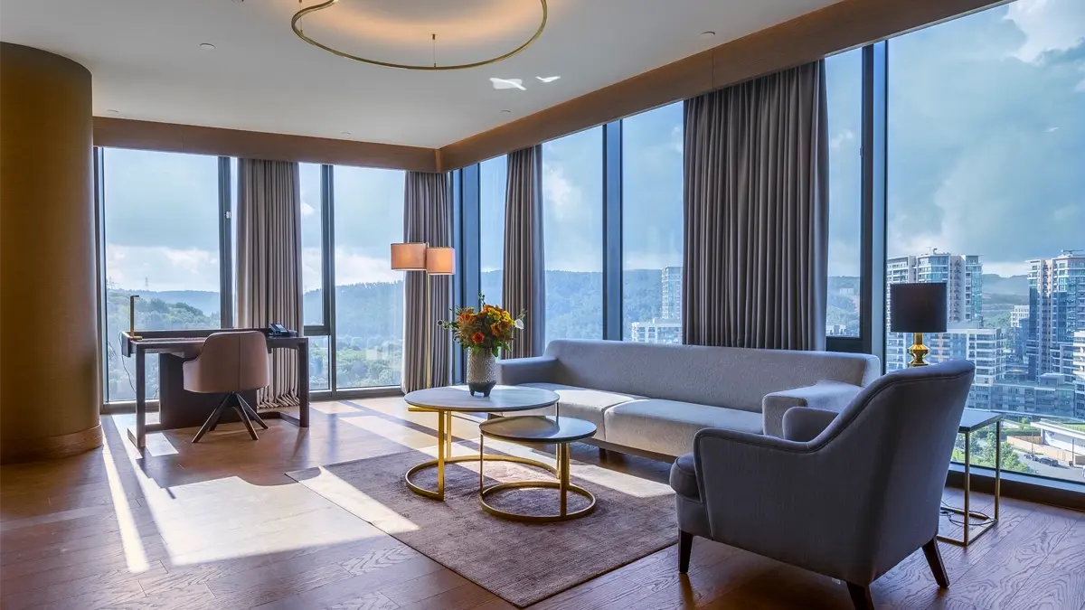 A sunny hotel room on an upper floor with a table and a few coaches and sofas and a 360 degree city view through French windows.